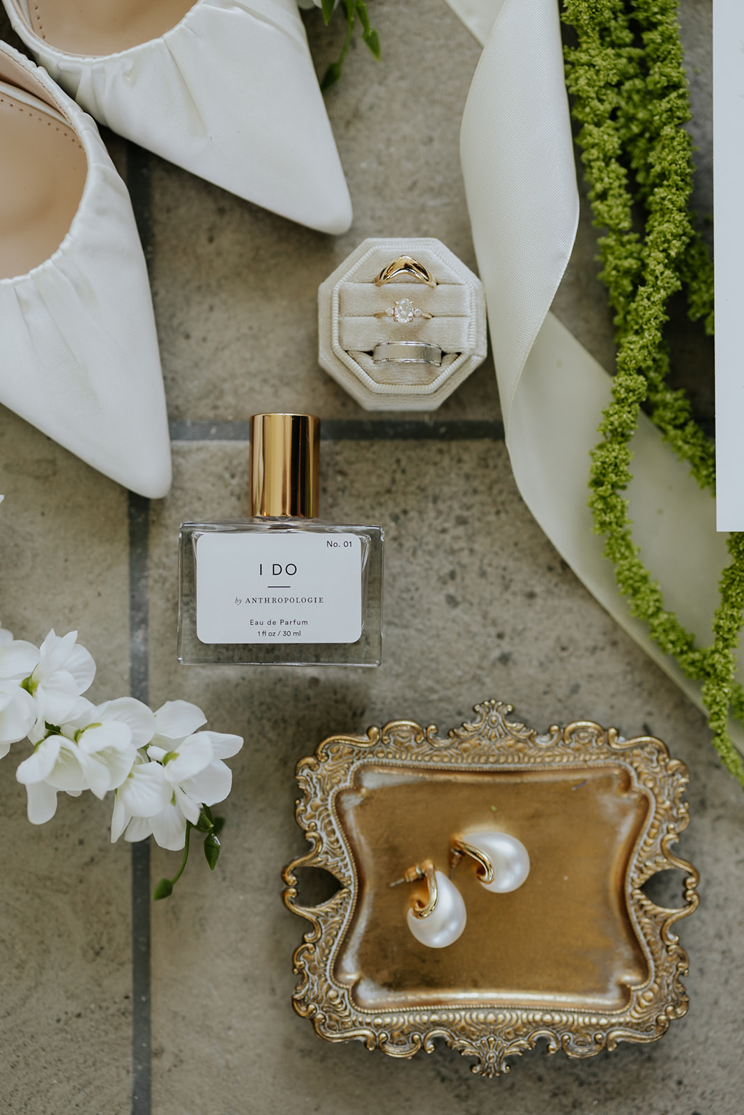 Wedding details: white heels, ring box, I Do perfume, pearl earrings on a gold tray and greenery.