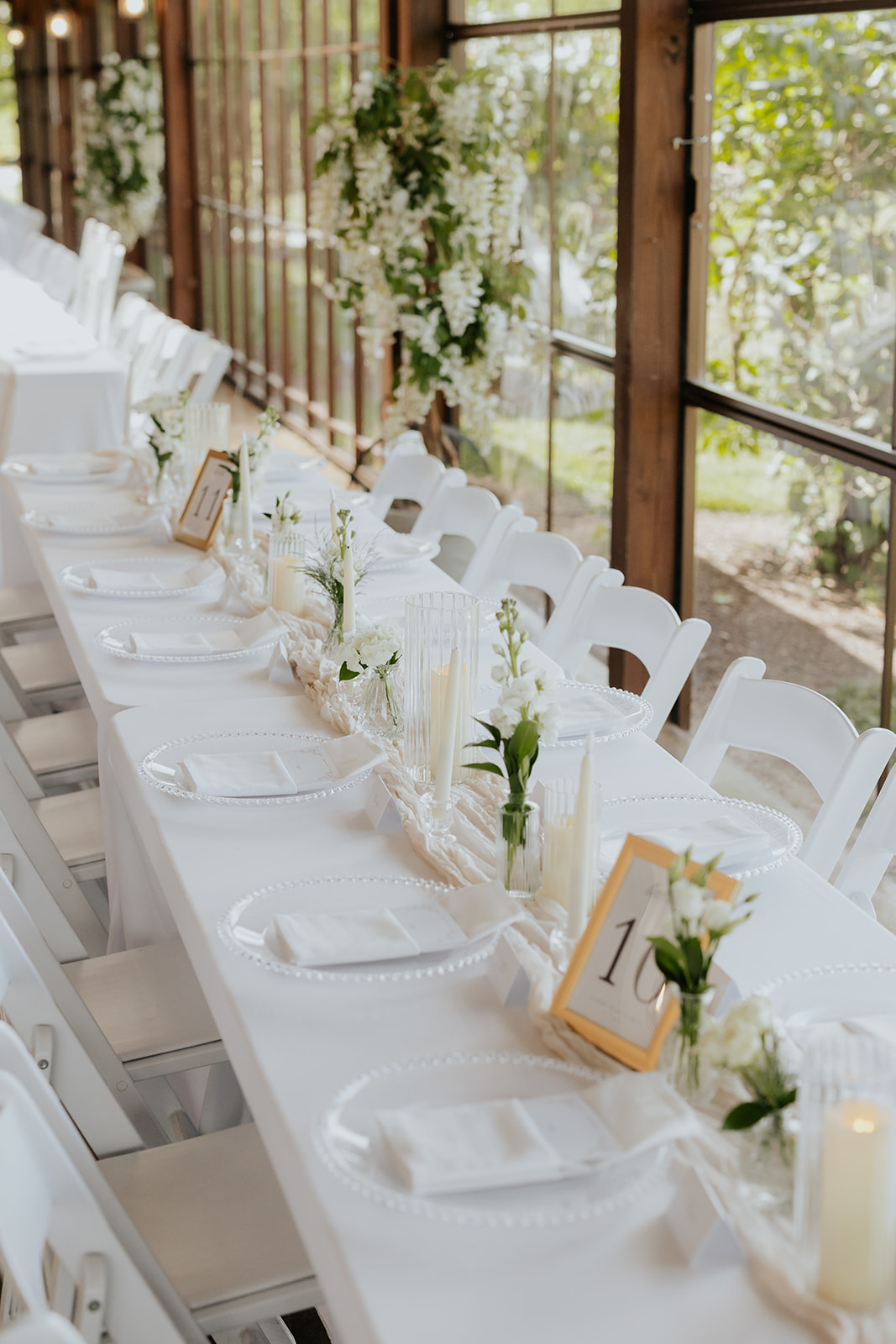 Long reception table set with white linens, beaded plates, candles, small bouquets and gold table numbers.
