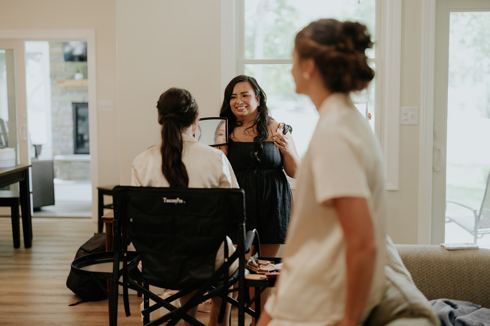 A bridesmaid holds up a hand mirror and smiles as the bride gets ready in her robe.