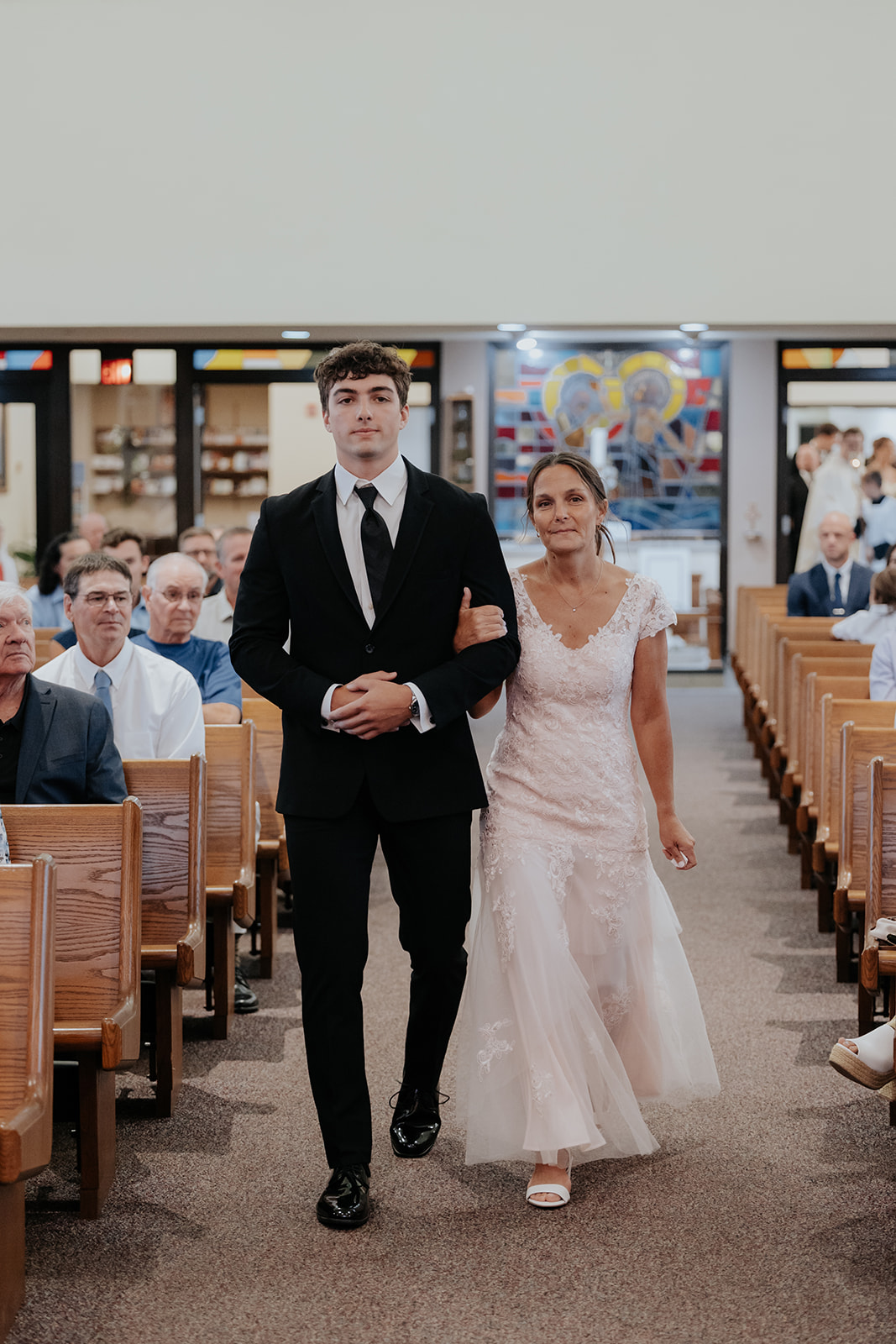 A young man in a black suit escorts a woman in a lace dress down the church aisle.