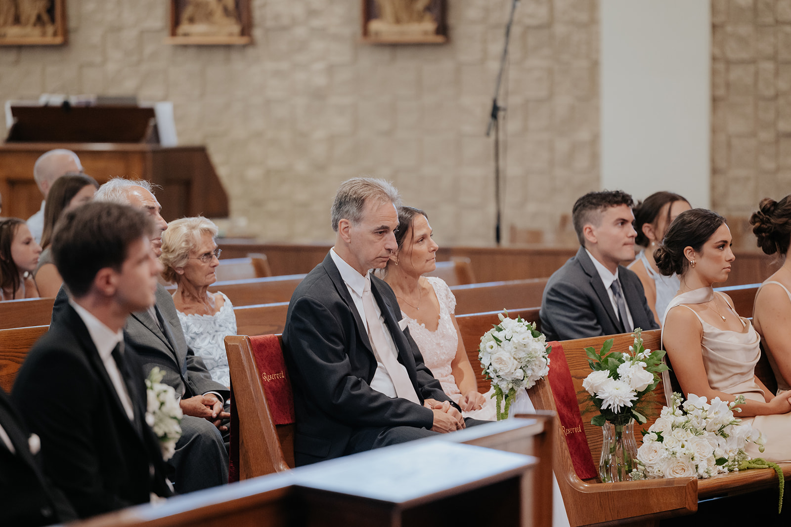 Family and bridesmaids sit in the front pews watching the church ceremony, bouquets beside them.