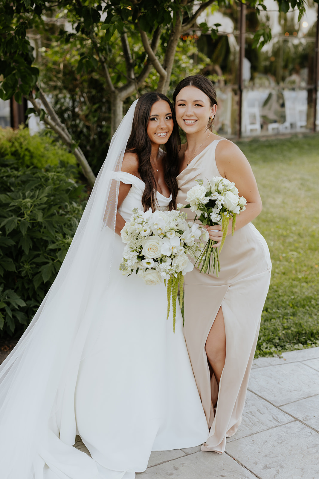 The bride and a bridesmaid in a champagne dress smile together holding white bouquets.