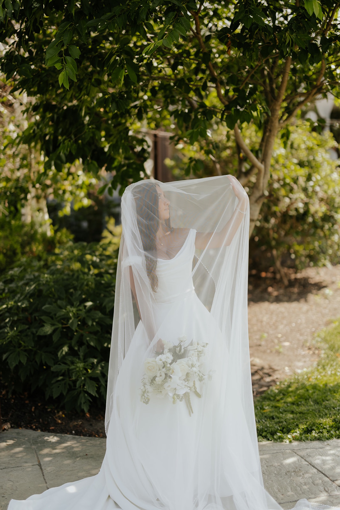 The bride lifts her long cathedral veil over her head beneath a shady tree, bouquet in hand.