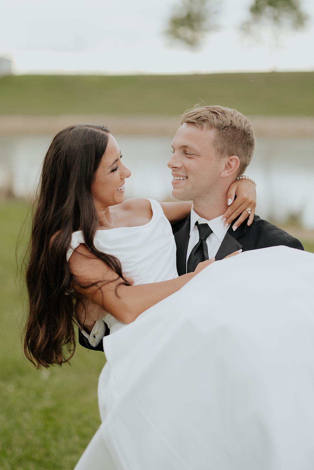Benjamin carries Isabel in his arms as they laugh together in front of a pond.