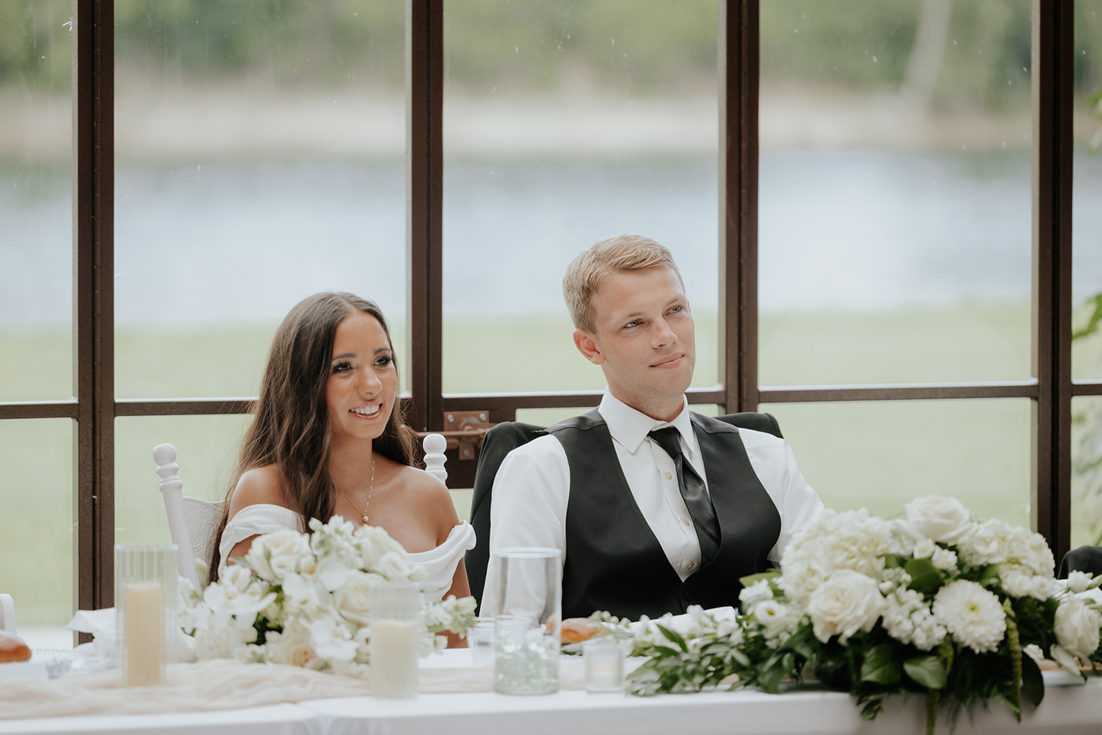Isabel and Benjamin smile from their sweetheart table during toasts, white flowers in front of them.