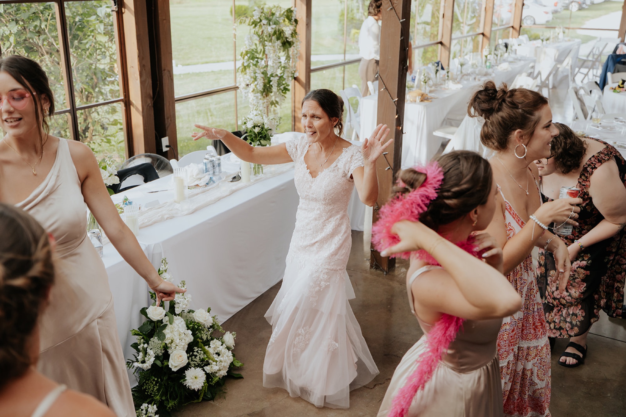 A woman in a lace wedding dress dances with bridesmaids and guests at the reception.