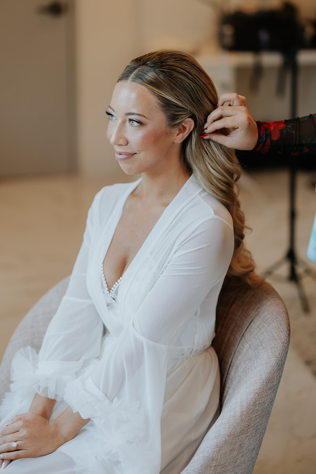 The bride smiles in a white robe as her stylist finishes her long curled hair