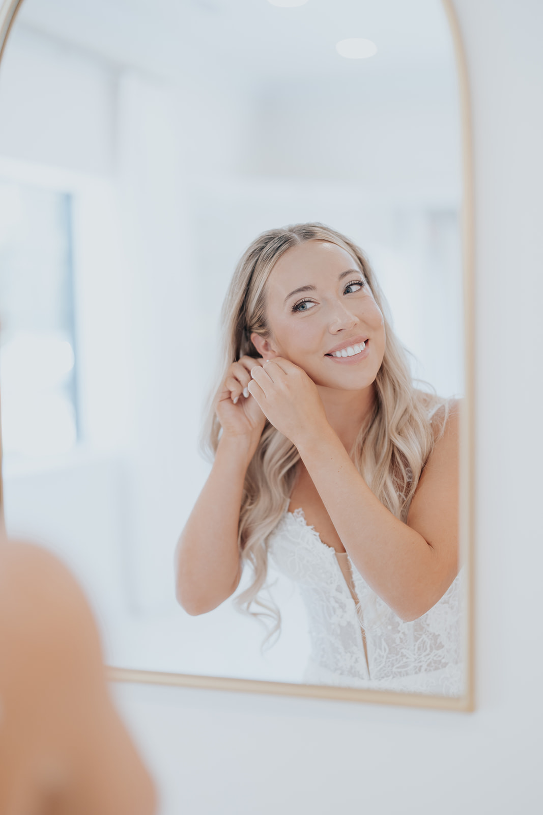 The bride smiles into an arched mirror as she puts on her pearl earrings