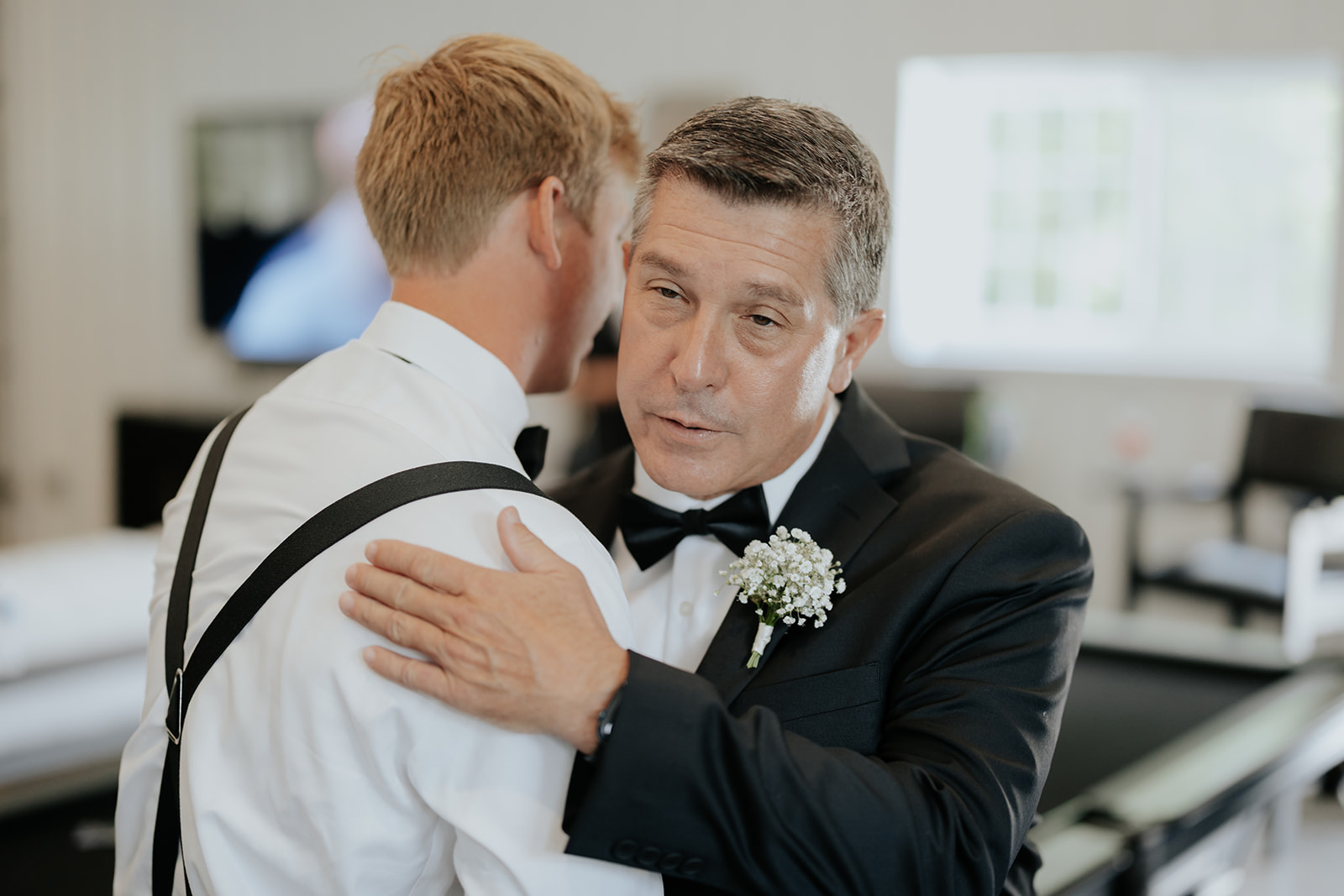 A man in a black tuxedo embraces the groom in suspenders while they get ready