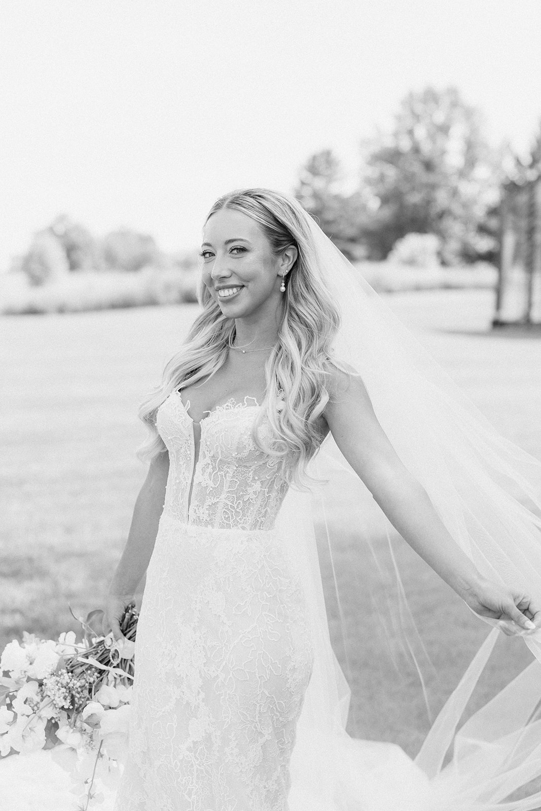 Black and white portrait of the bride smiling as she lifts her long veil in an open field