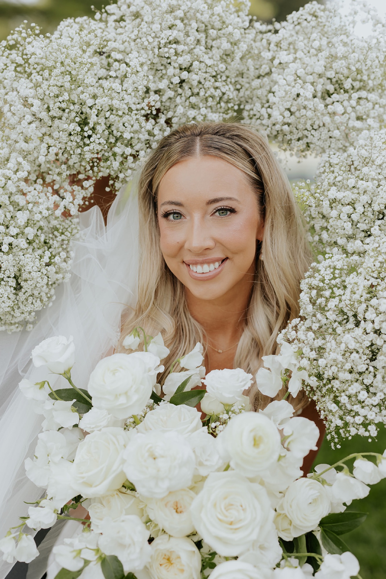 The bride smiles inside an arch of baby's breath, holding a bouquet of white roses