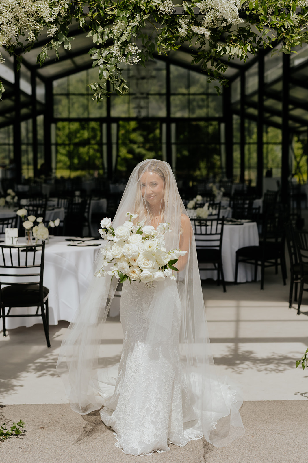 The bride stands beneath her veil holding white roses in the glass reception pavilion