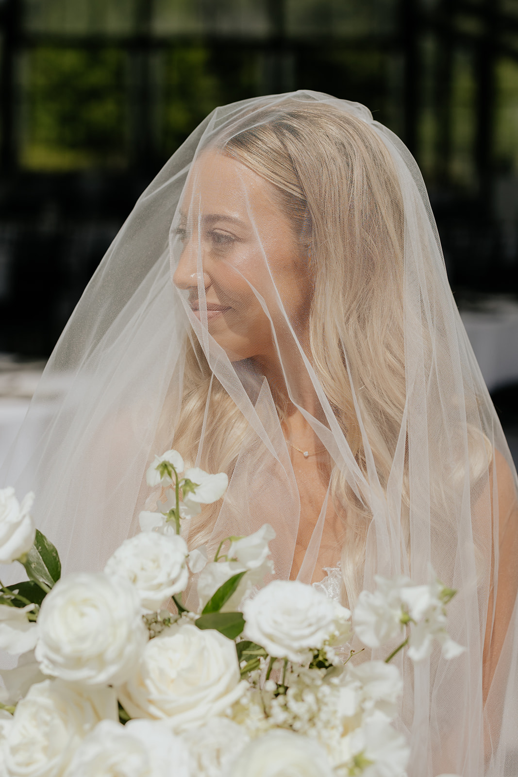 The bride smiles softly through her veil, a bouquet of white roses in the foreground