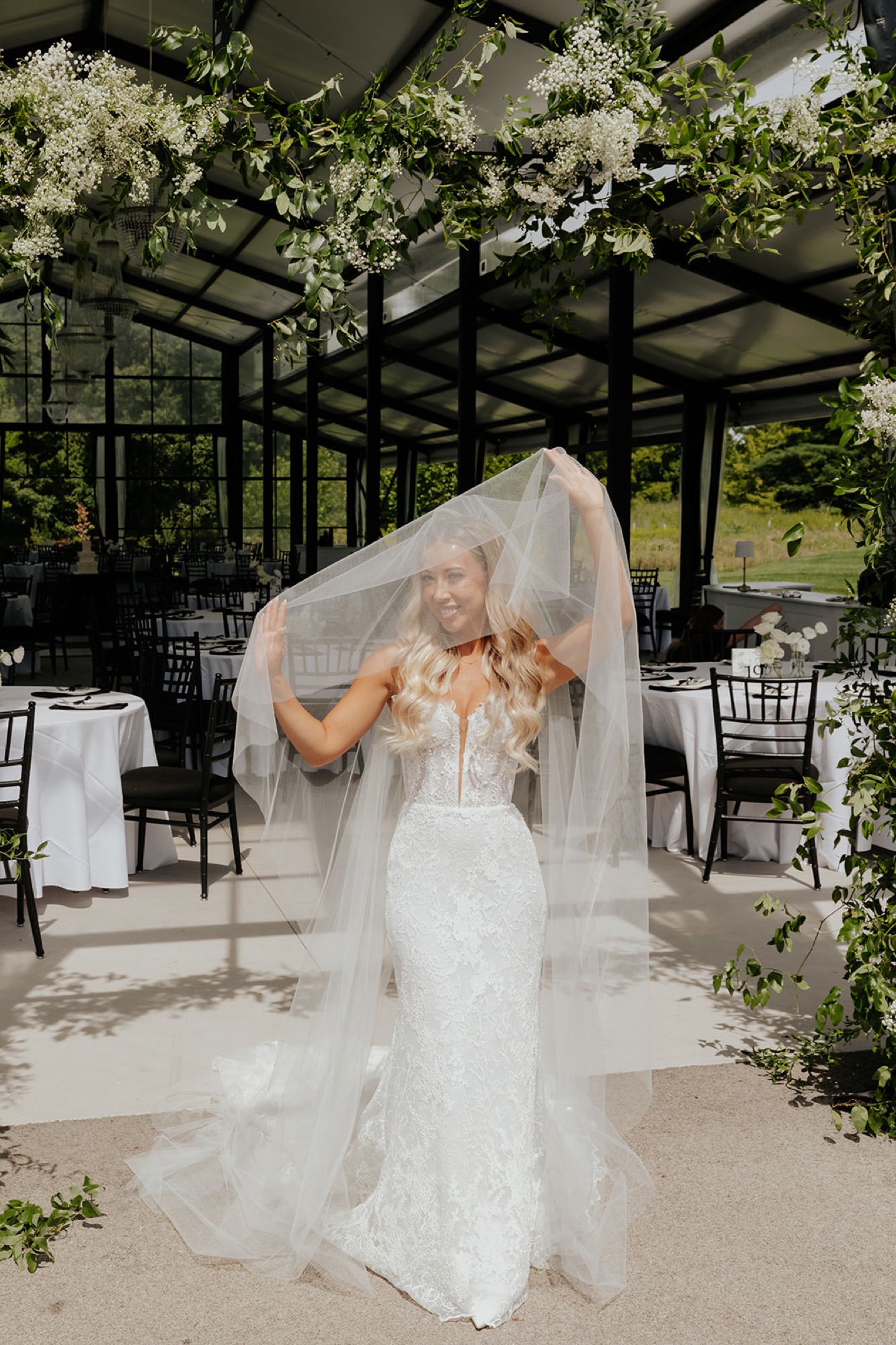 The bride holds her veil overhead beneath hanging greenery and white flowers in the pavilion