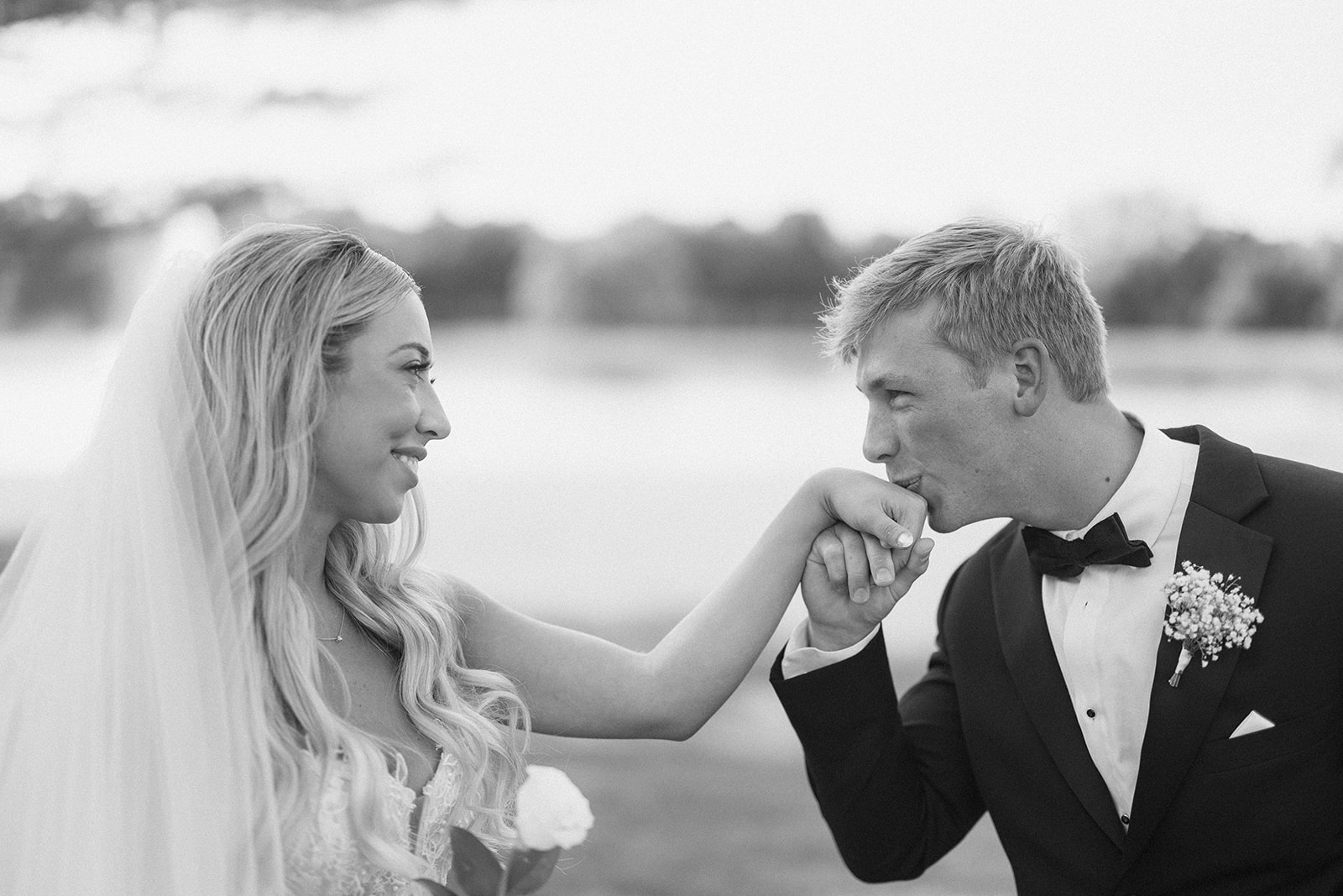 Black and white photo of Grant kissing Lauren's hand as she smiles beside the water