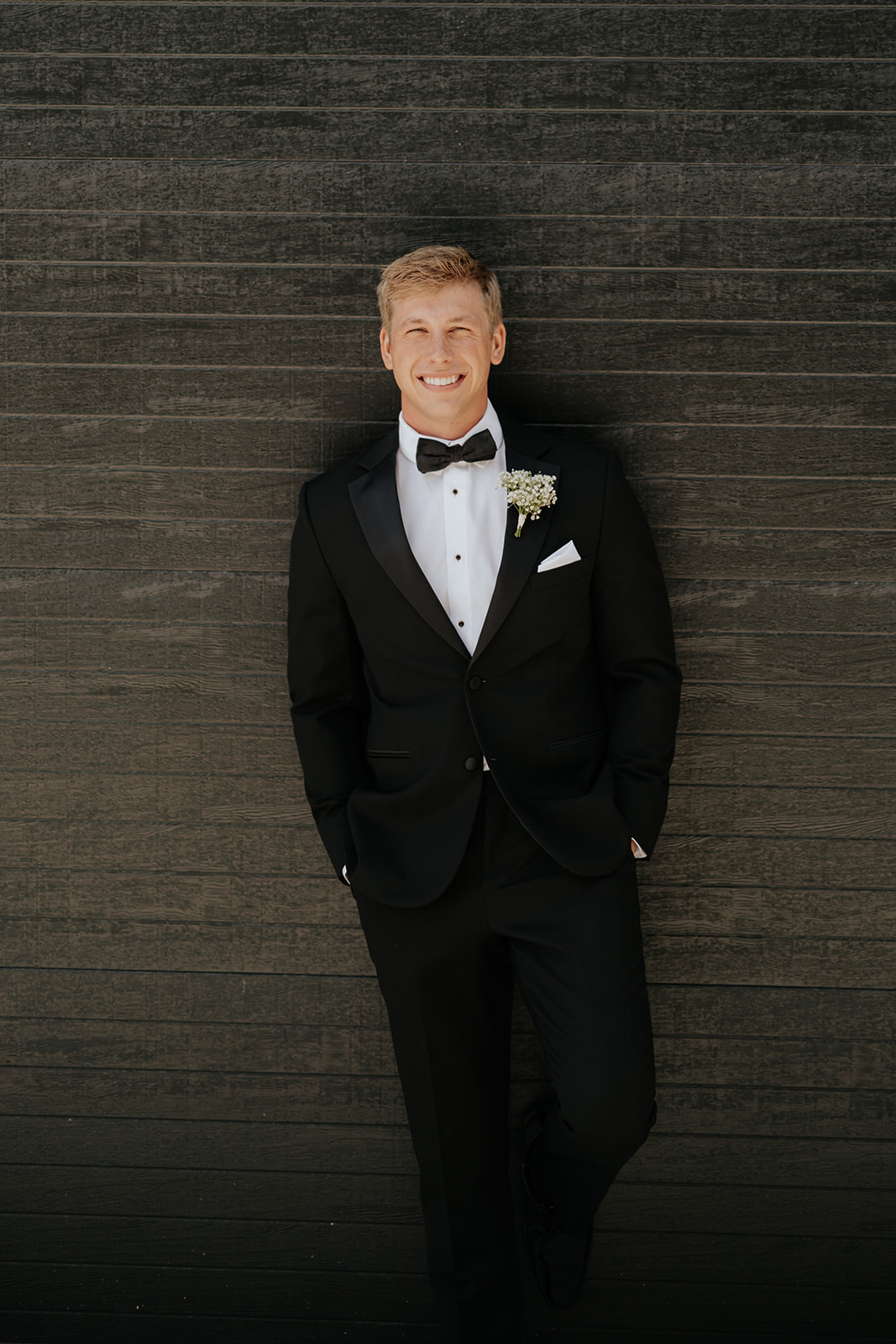 The groom leans against a dark wood wall in a black tuxedo with a baby's breath boutonniere