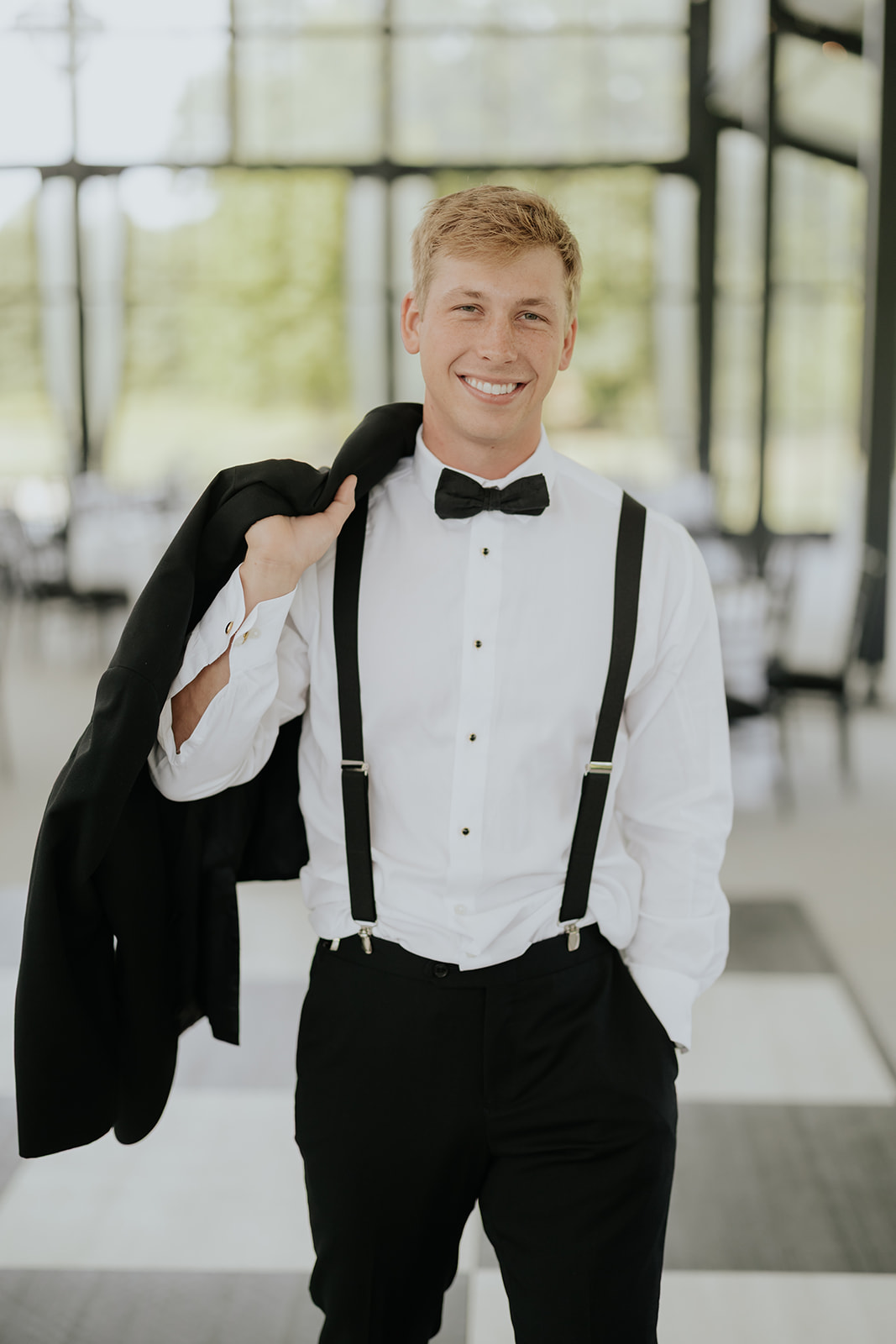 The groom smiles with his tuxedo jacket slung over his shoulder in suspenders and bow tie