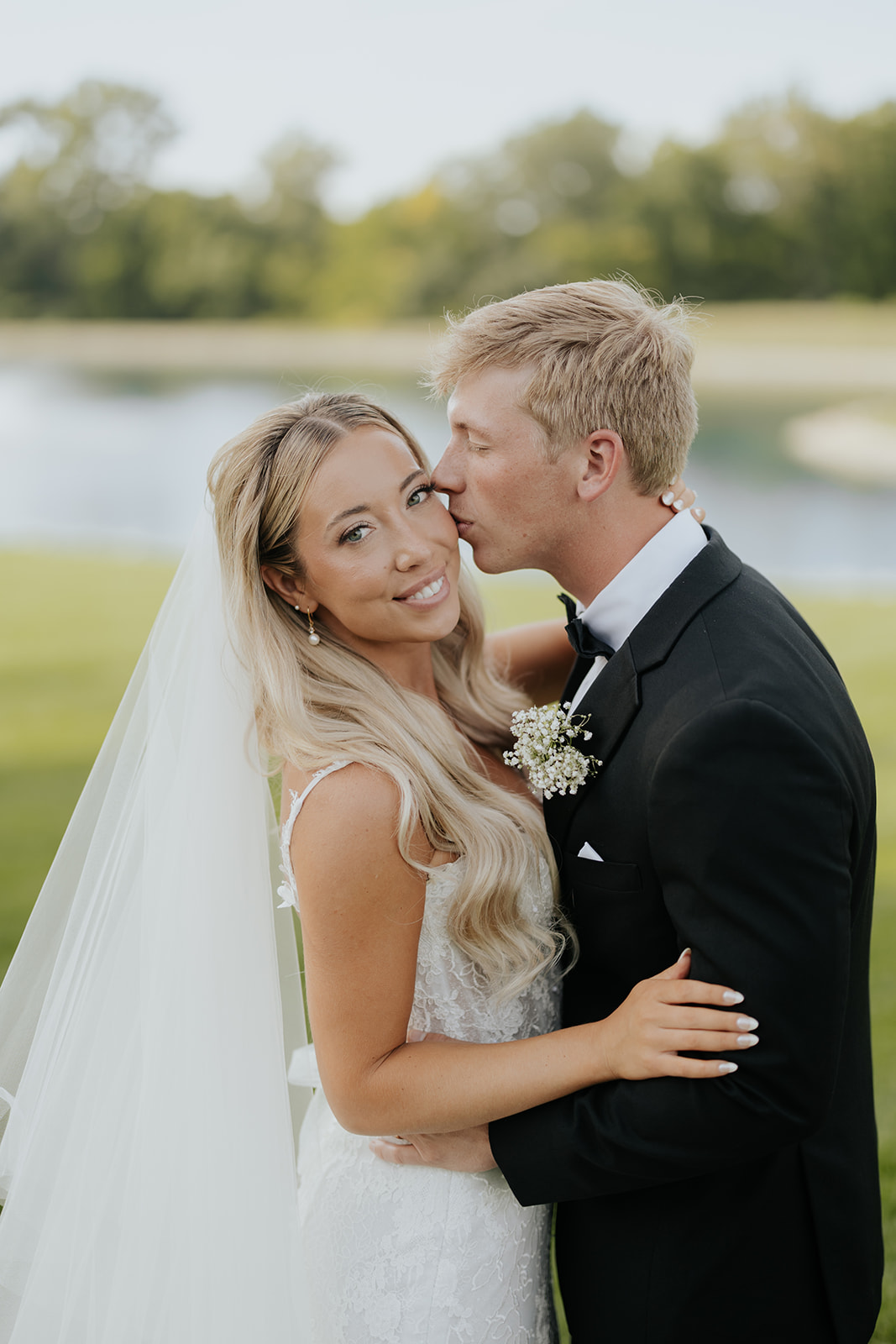 Grant kisses Lauren's cheek as she smiles, a pond and trees behind them