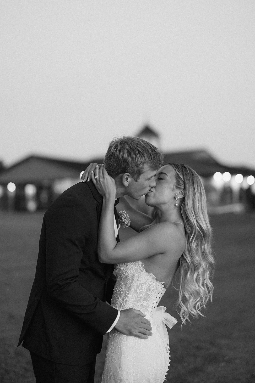 Black and white photo of Lauren and Grant kissing at dusk, the lit venue behind them