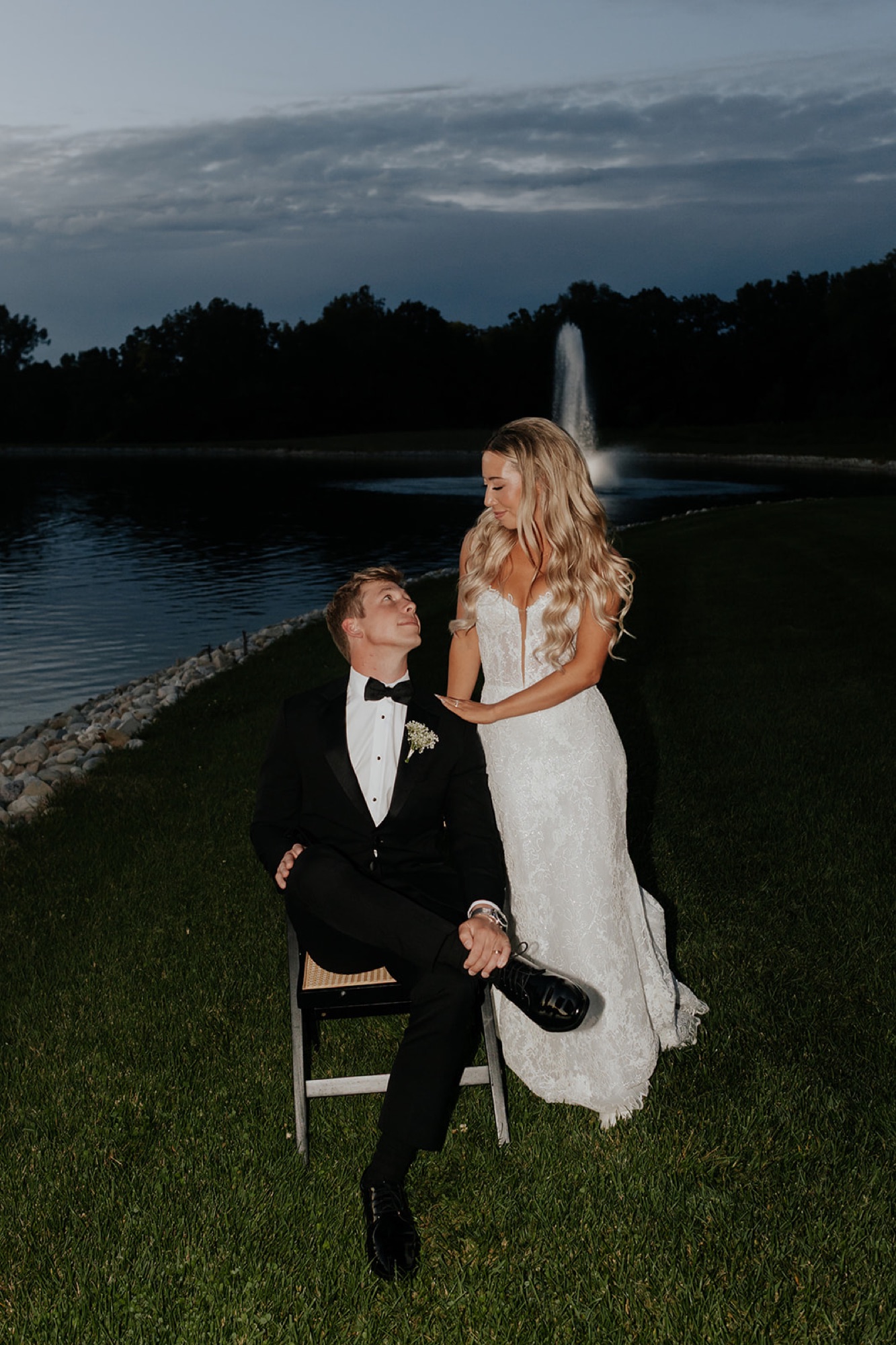 The groom sits on a chair as the bride rests on his knee beside a lit fountain at dusk