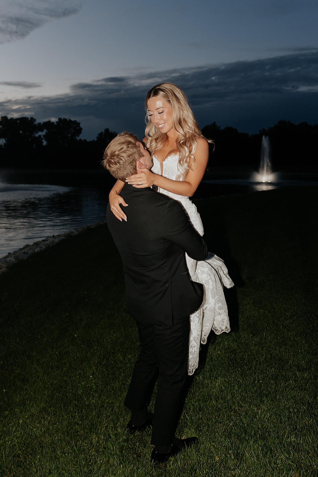Grant lifts Lauren off the ground at dusk beside the pond with the fountain glowing behind