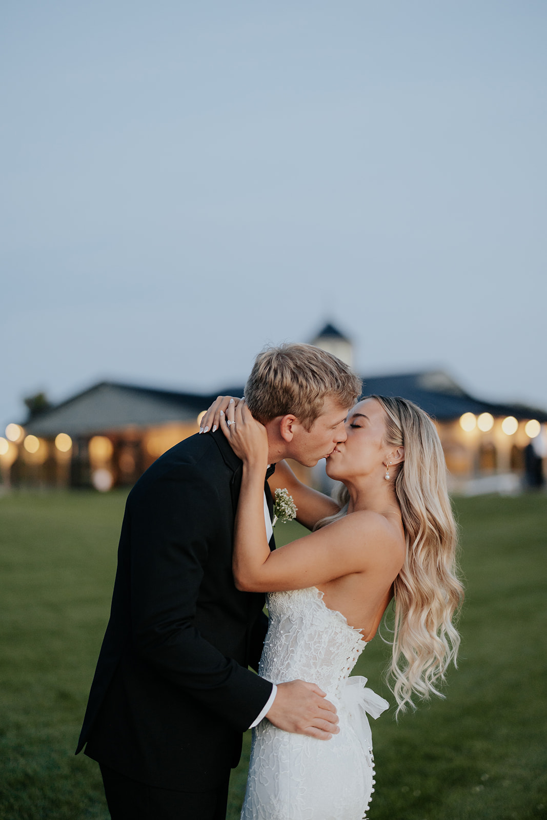 Lauren and Grant kiss on the lawn at dusk with string lights glowing on the venue behind