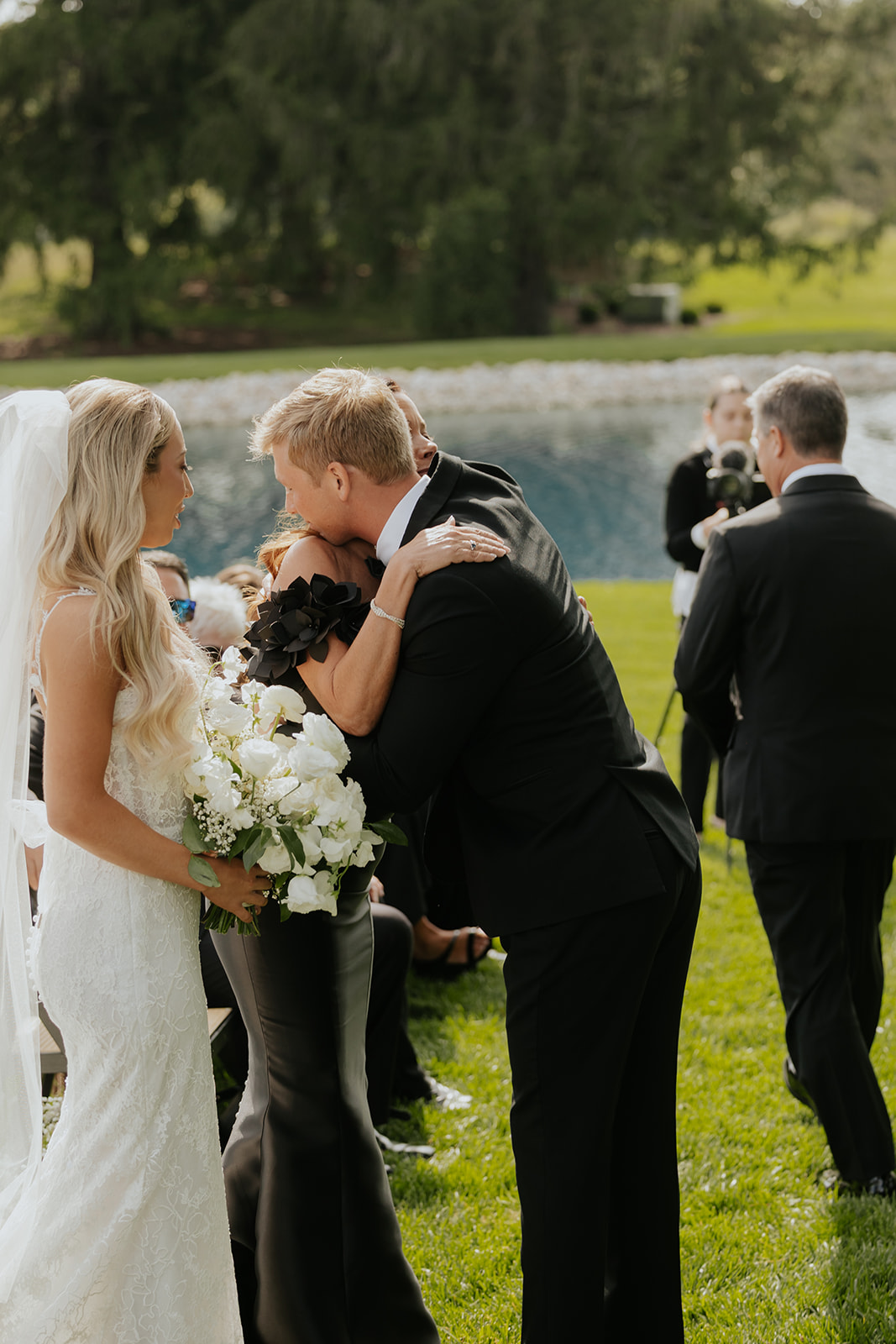 The groom hugs a guest along the aisle while the bride looks on holding white roses