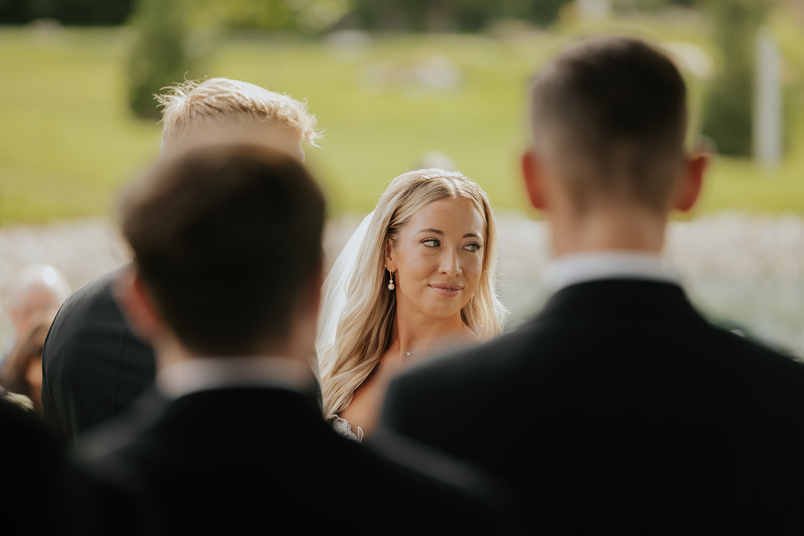 The bride smiles at the groom during the ceremony, framed between groomsmen's shoulders
