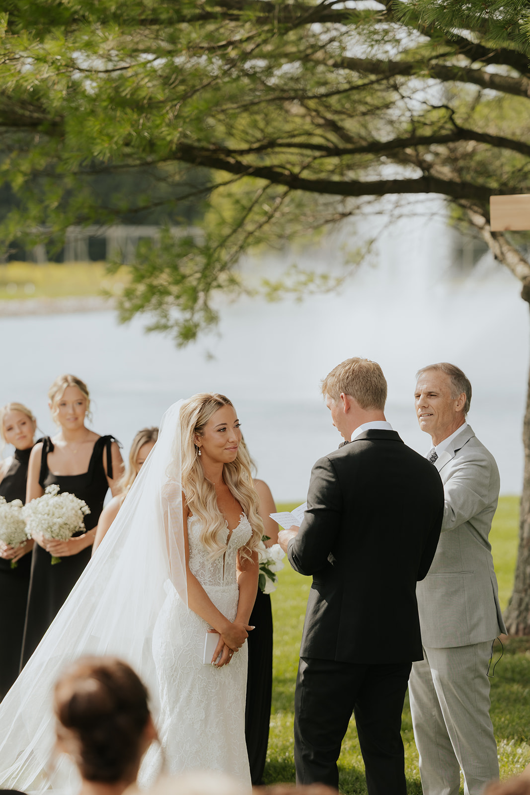 Lauren and Grant hold hands at the altar under a tree beside the pond, bridesmaids looking on