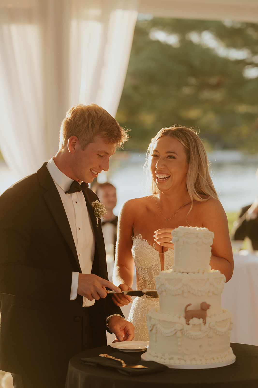 Lauren and Grant laugh while cutting their white tiered cake in golden evening light