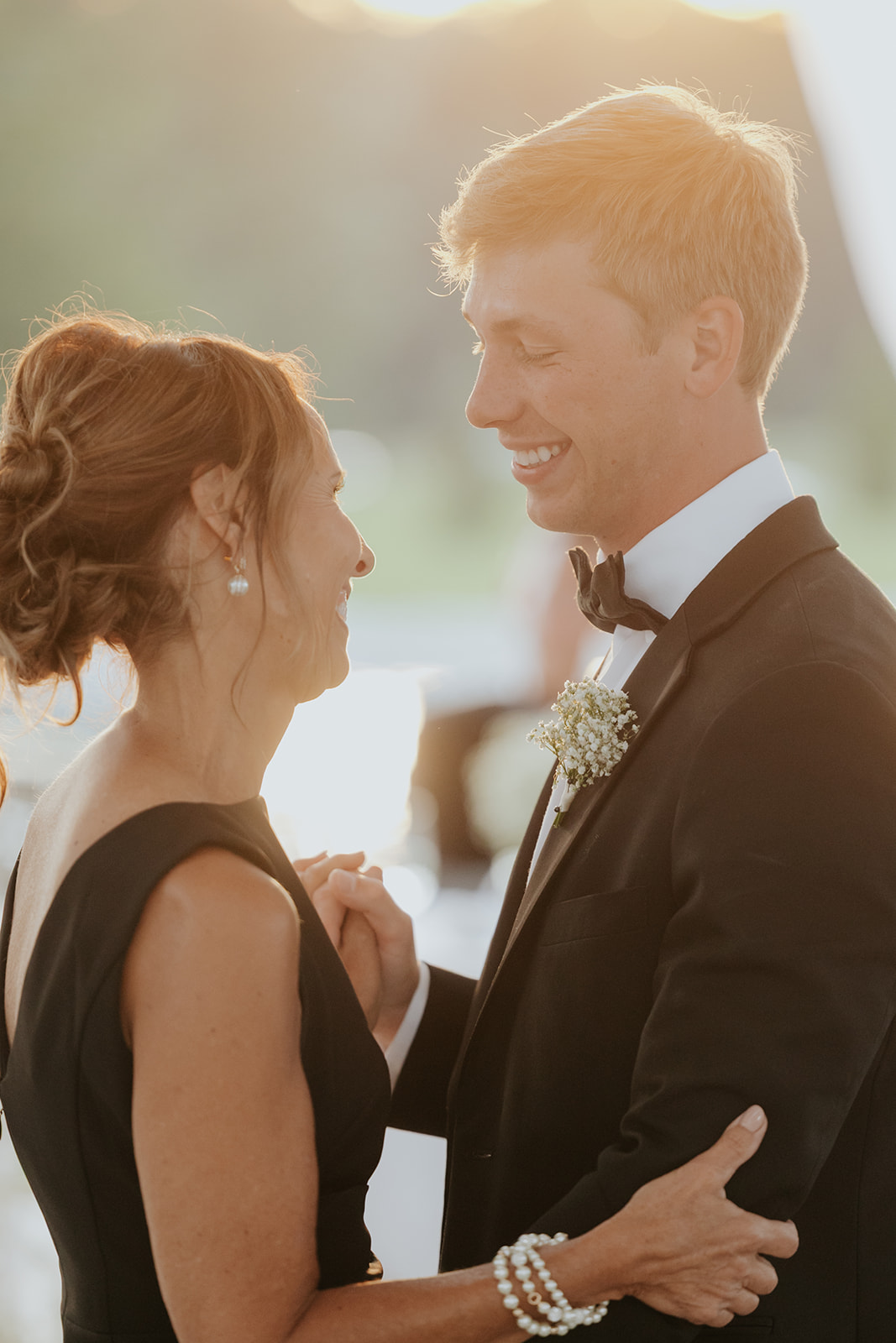 The groom shares a smiling dance with a woman in a black dress in golden evening light