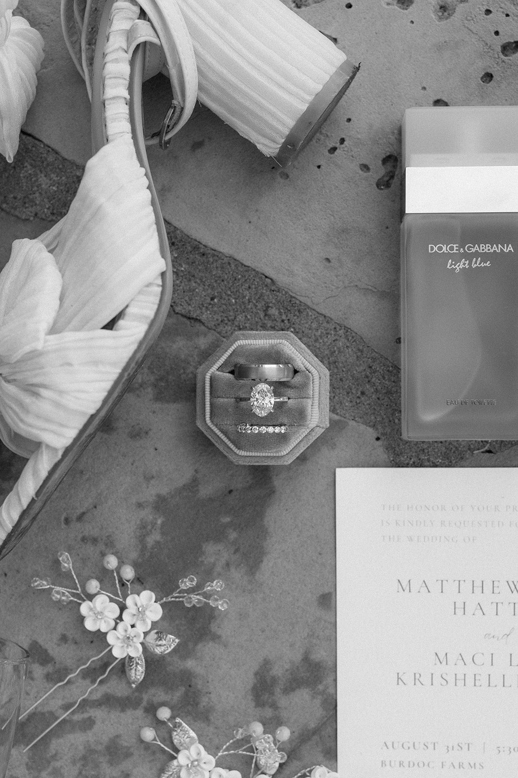 Black and white flat lay of the engagement ring, invitation, perfume, hair pins and bridal heels
