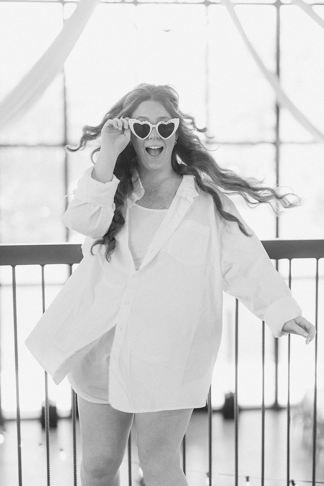 Black and white of the bride in heart sunglasses and a button-up shirt, laughing on a balcony