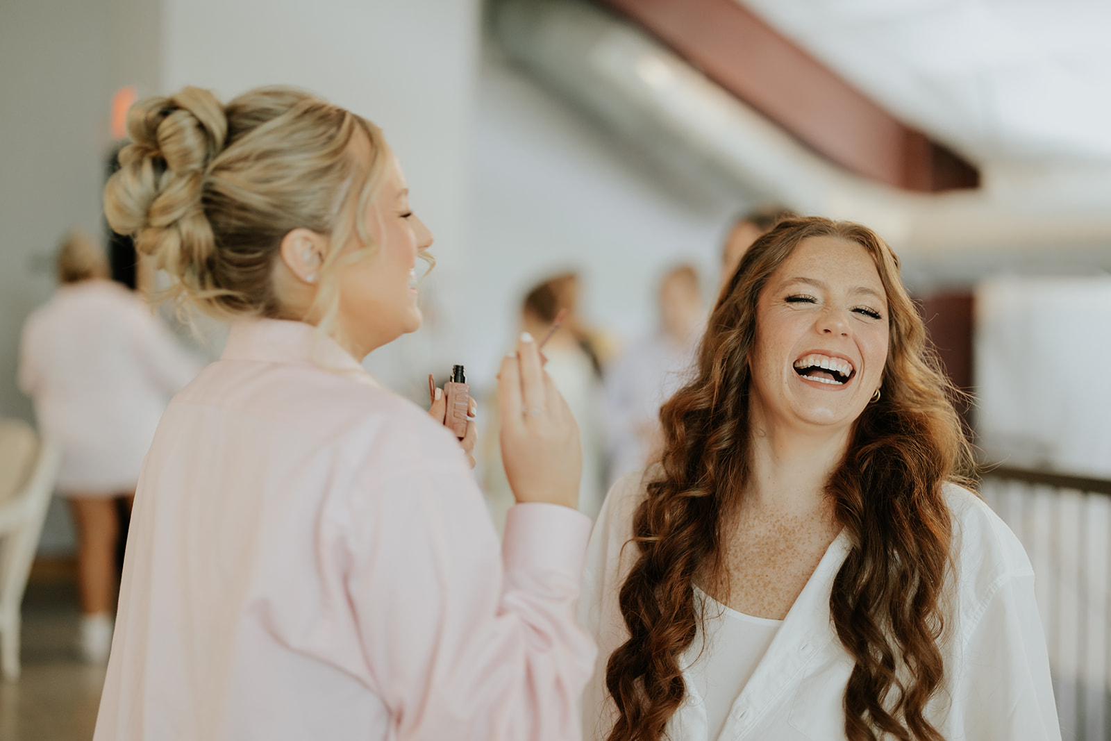 The bride laughs with a bridesmaid in a pink robe while getting ready