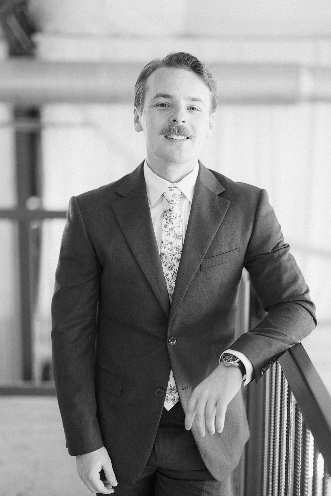 Black and white portrait of the groom leaning on a railing in his suit and floral tie
