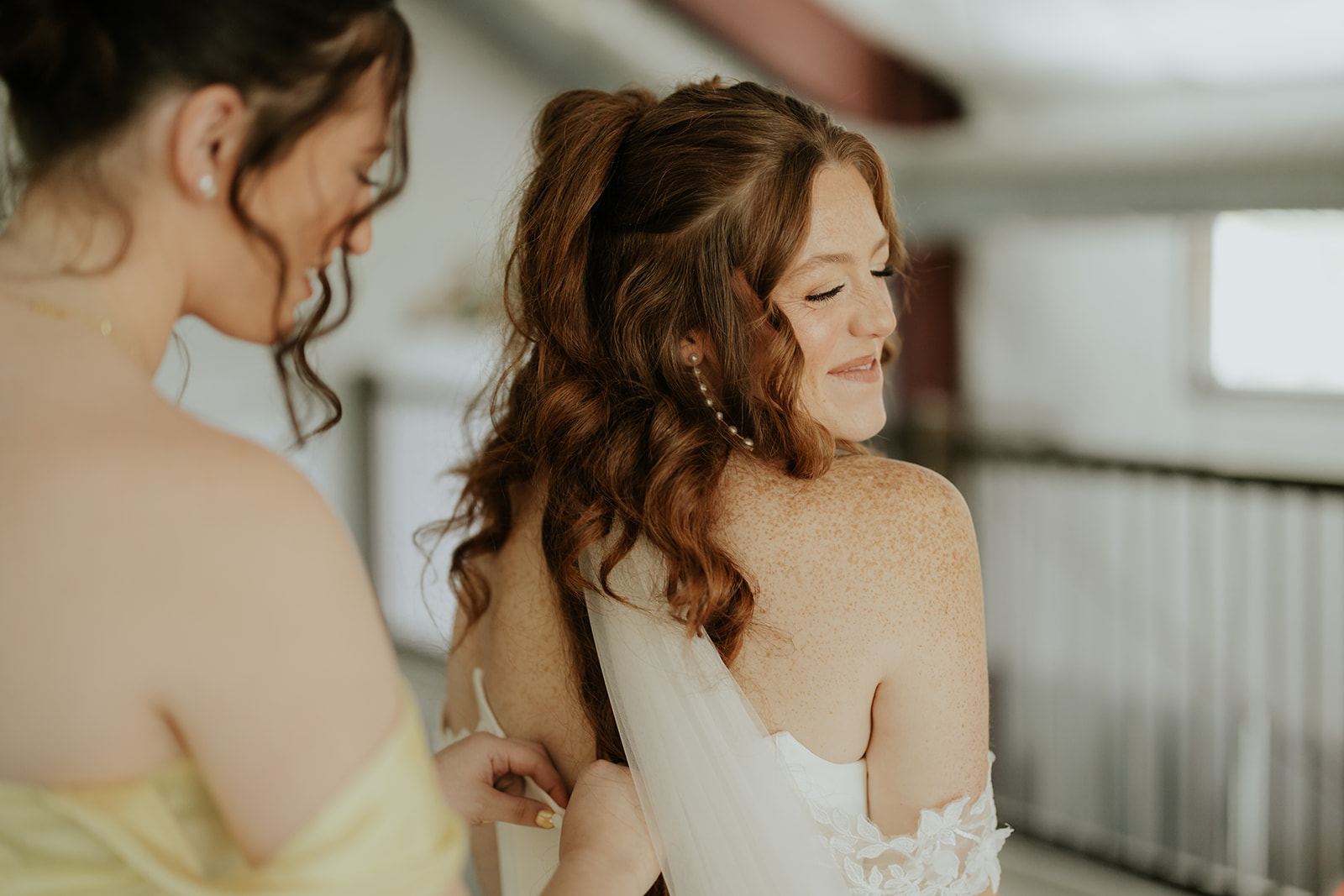 A bridesmaid in yellow fastens the back of the bride's off-the-shoulder gown