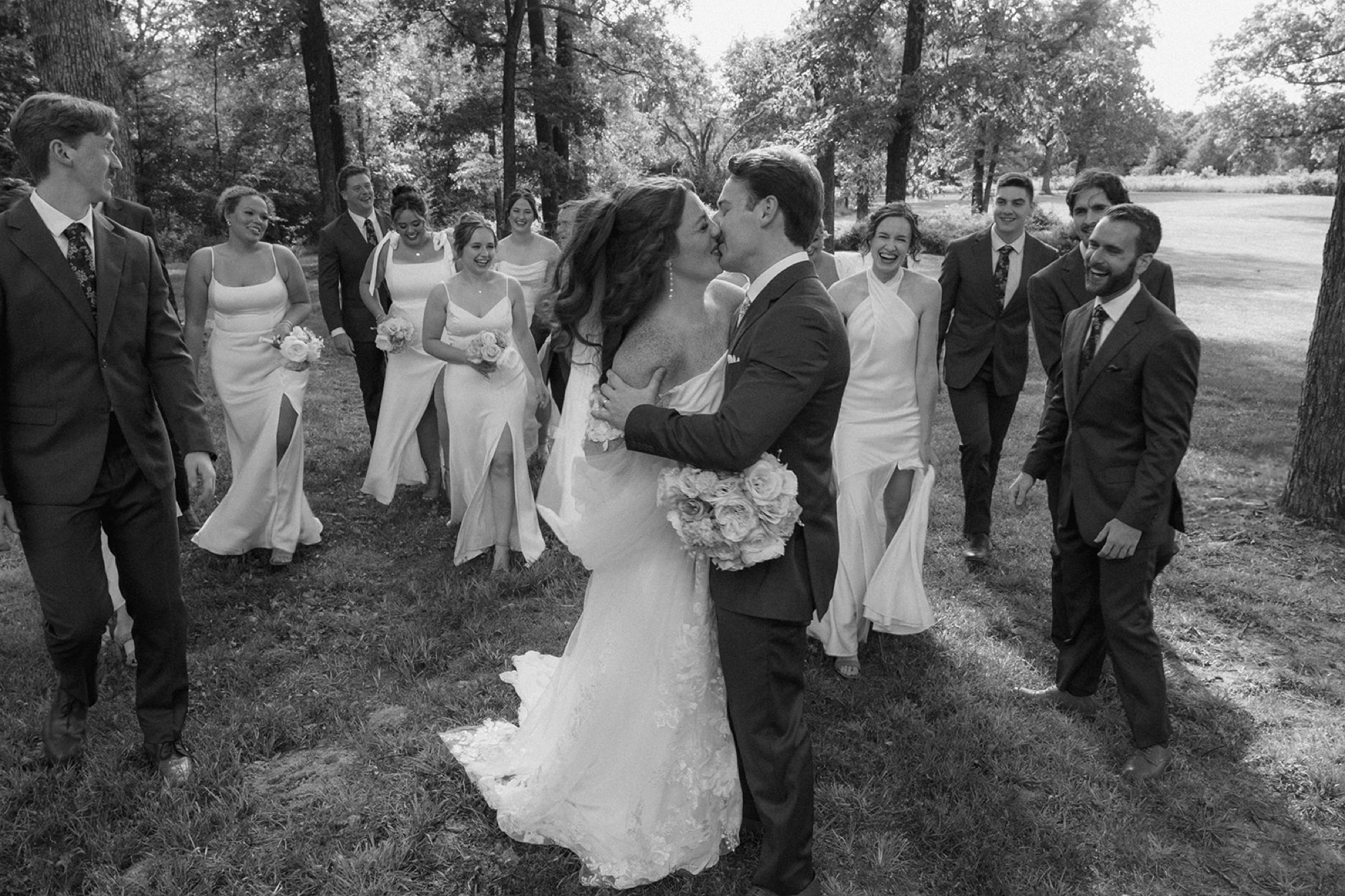 Black and white of Maci and Matt kissing as their wedding party walks behind them among the trees