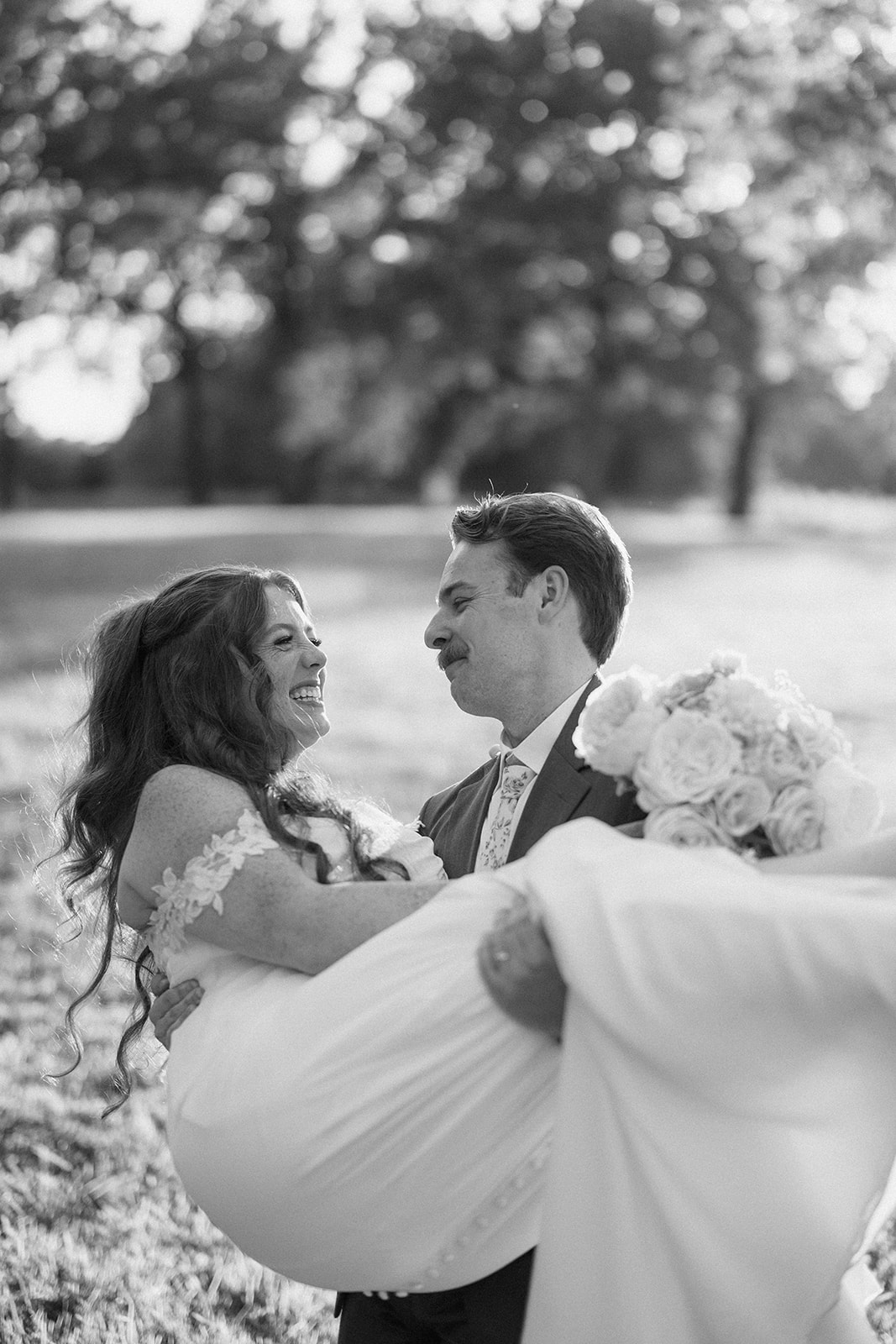 Black and white of the groom carrying the bride across the field as they both laugh