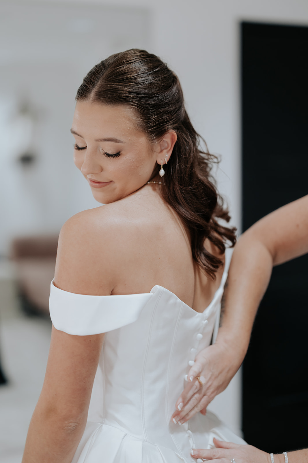 The bride smiles down as someone fastens the back of her off-the-shoulder gown