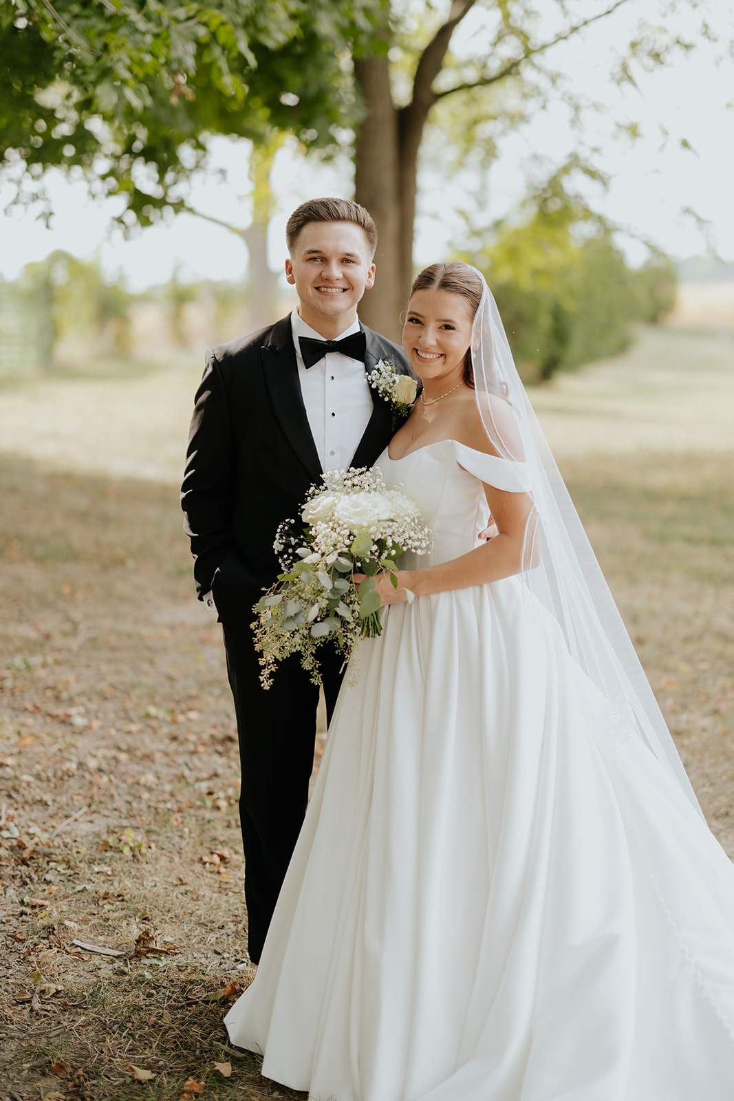 Regan and Reyn smile together on a tree-lined path, her holding a white bouquet