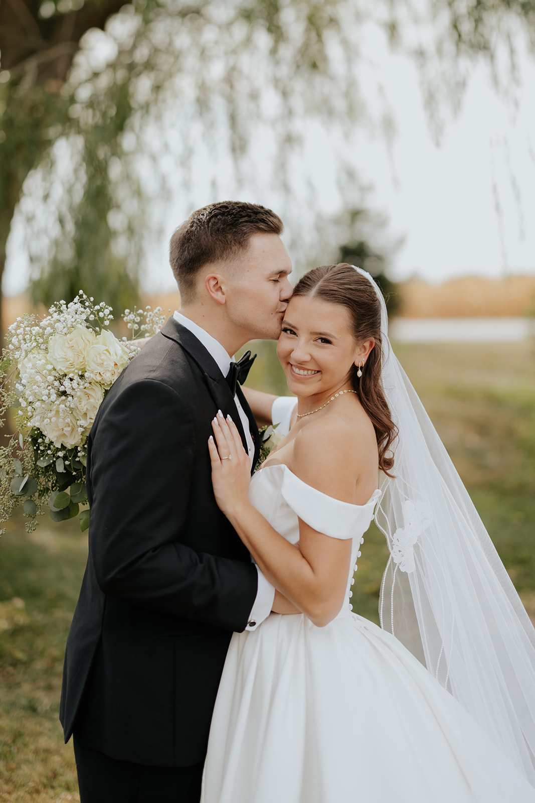 The groom kisses the bride's temple as she smiles, holding her bouquet