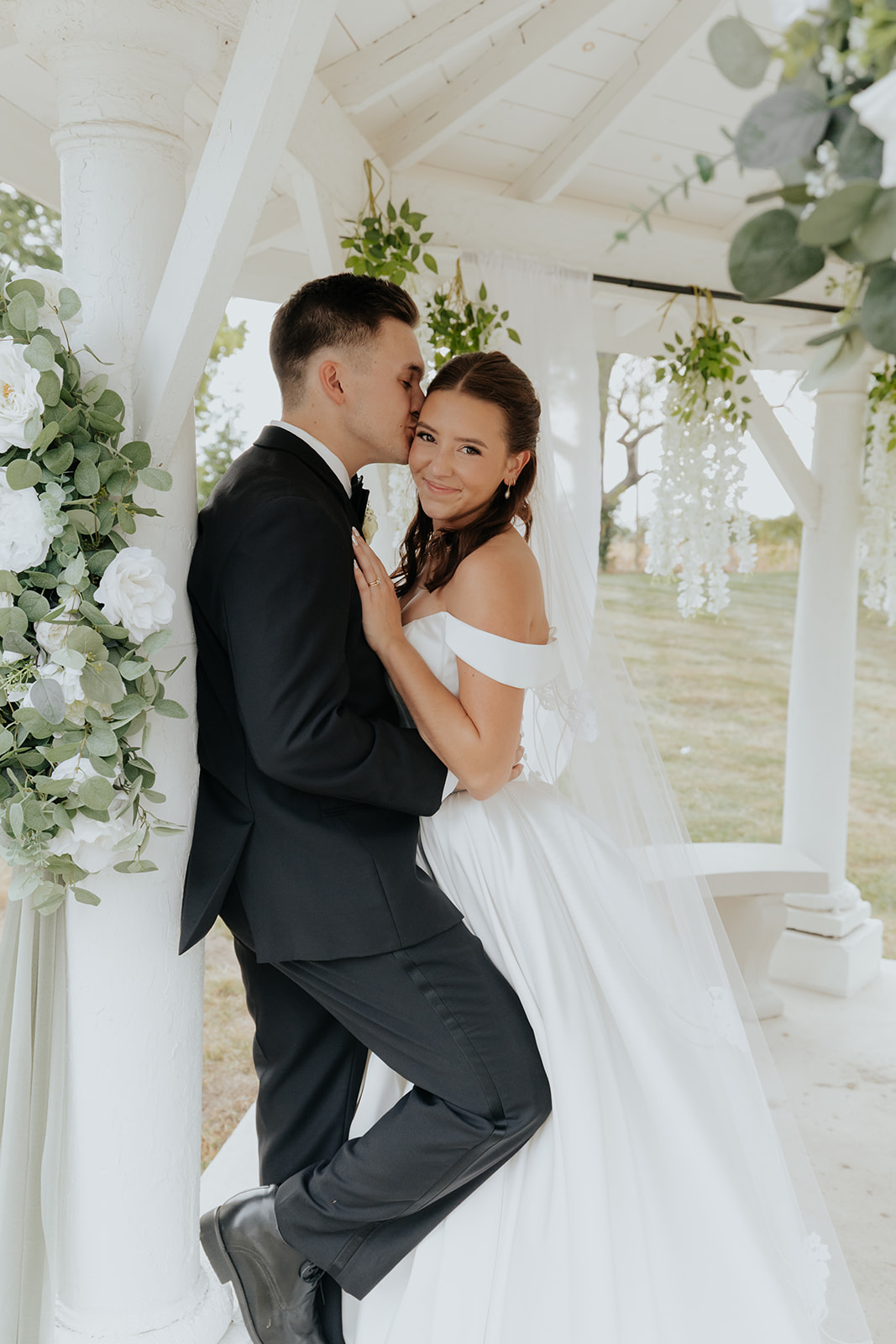 The groom kisses the bride's cheek as they lean against the white gazebo